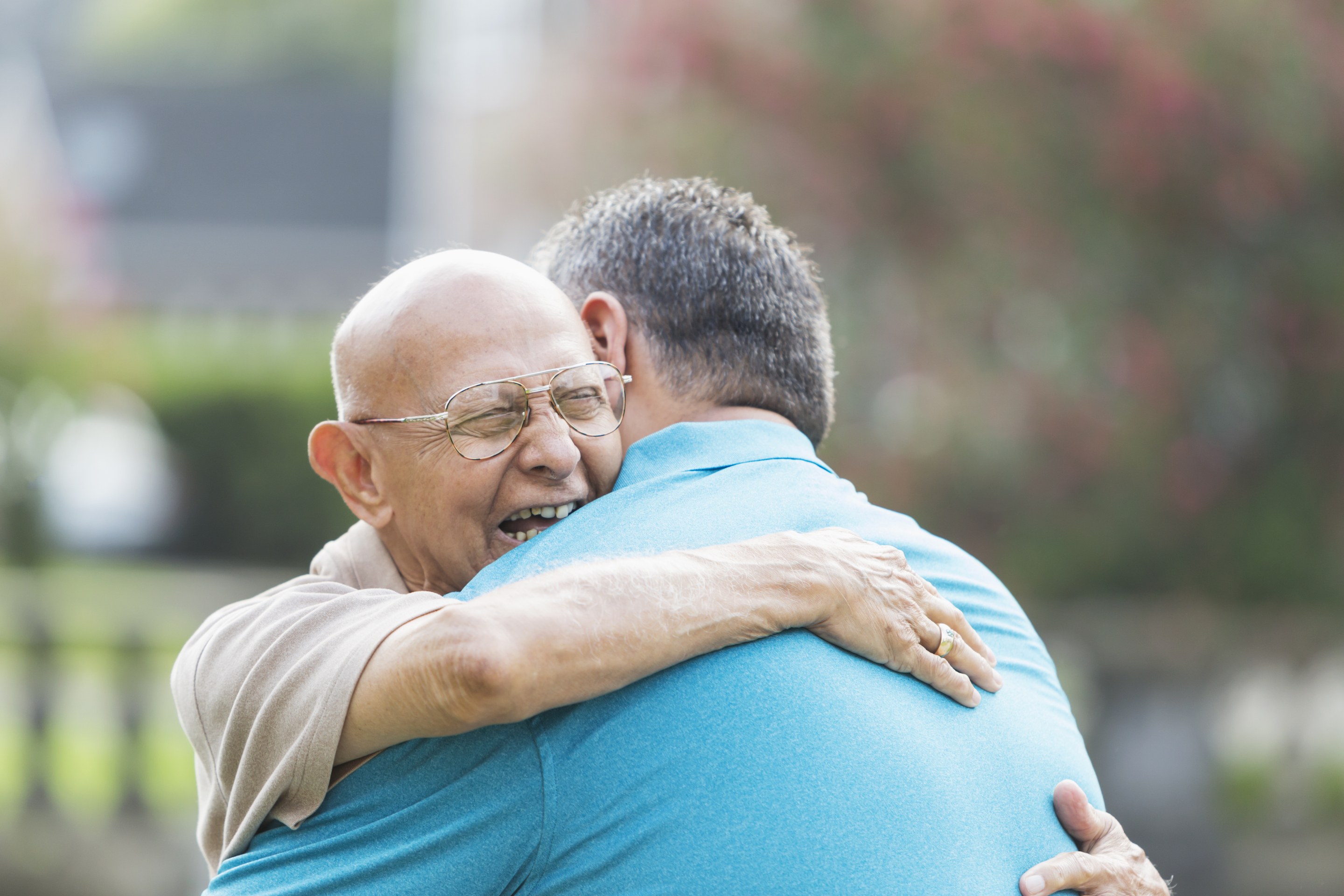 Elderly man with glasses hugging younger man in blue shirt outdoors