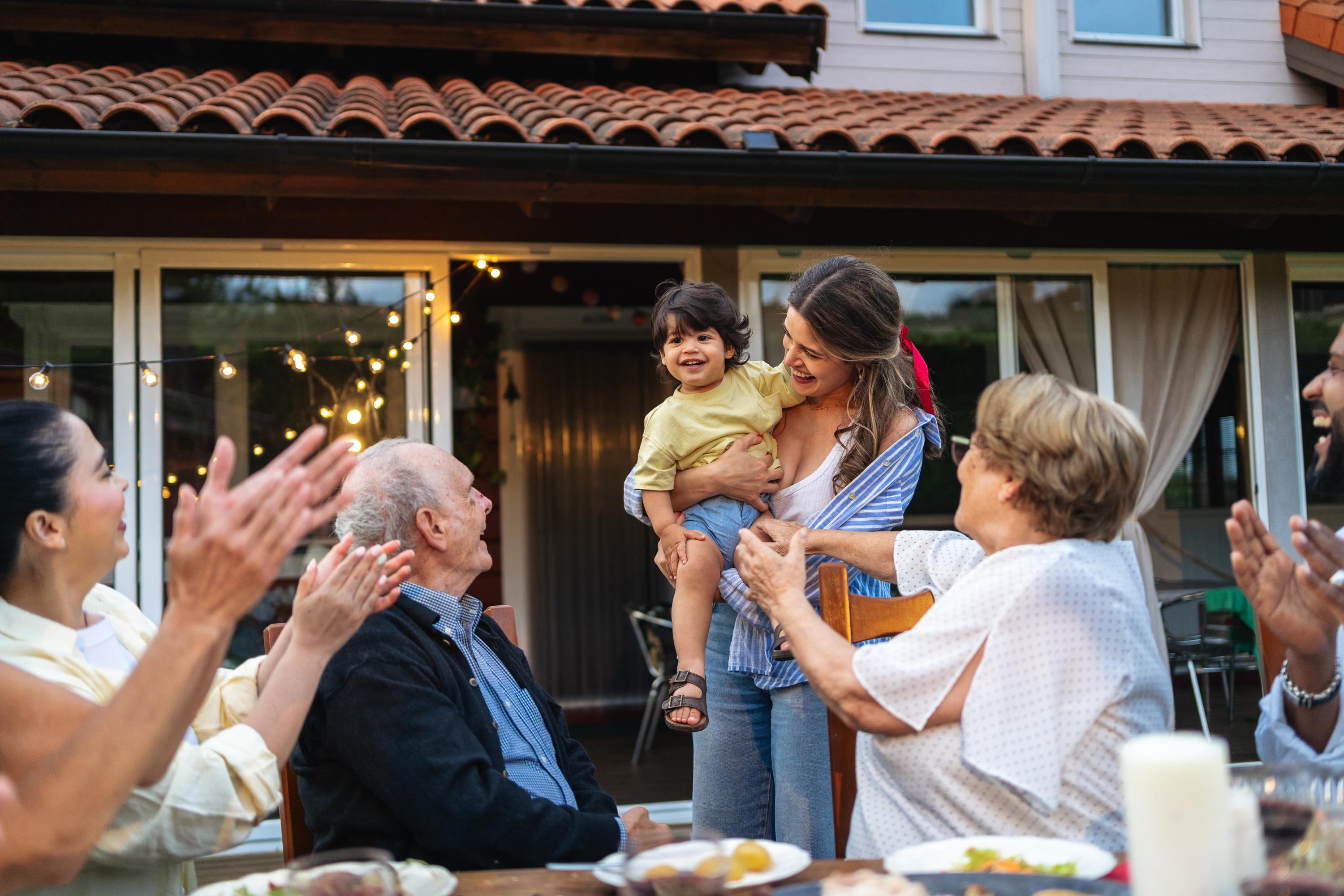 Family gathering with mother holding child while relatives applaud at outdoor dinner