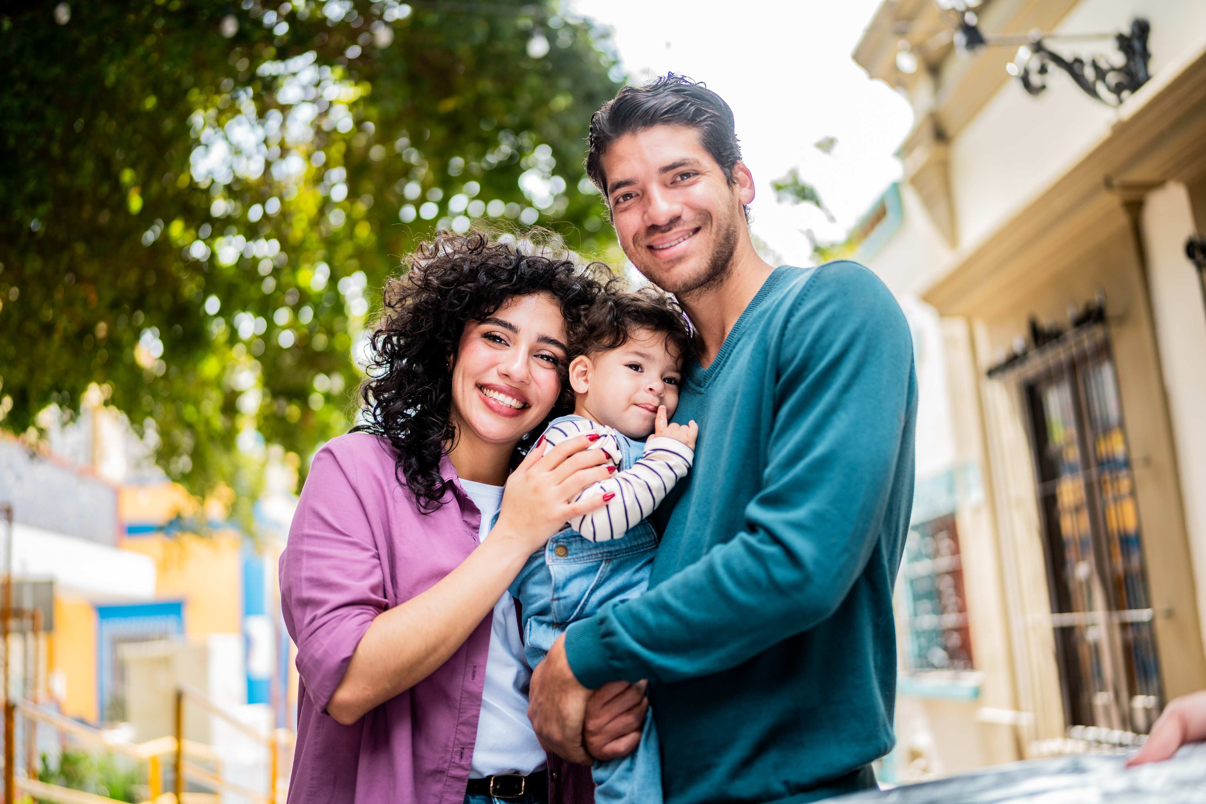 Happy family of three with curly haired mother in purple shirt, father in teal shirt, and young child in striped shirt standing together on a sunny street with trees and buildings in the background