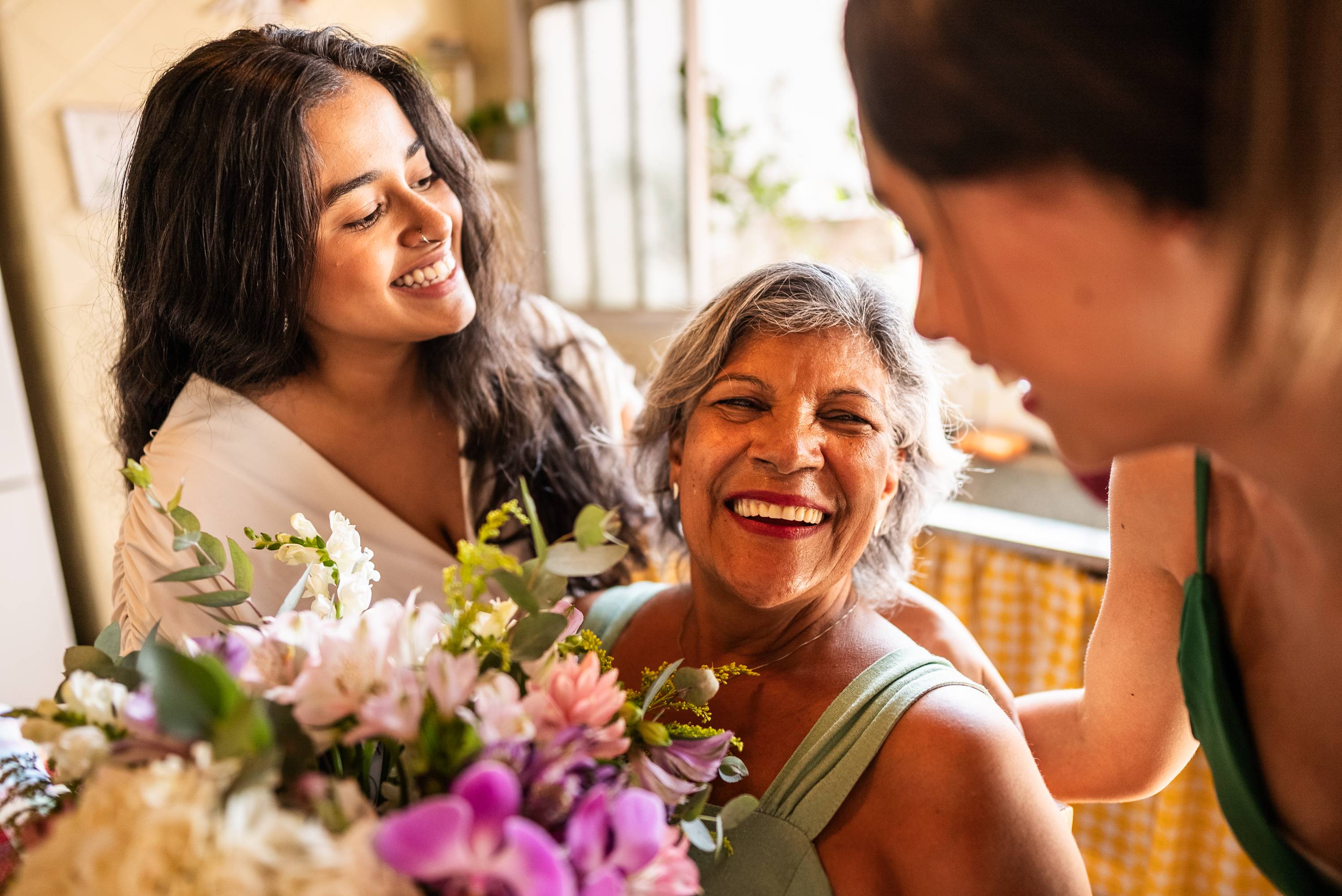 Three women smiling together with flowers in warm indoor setting