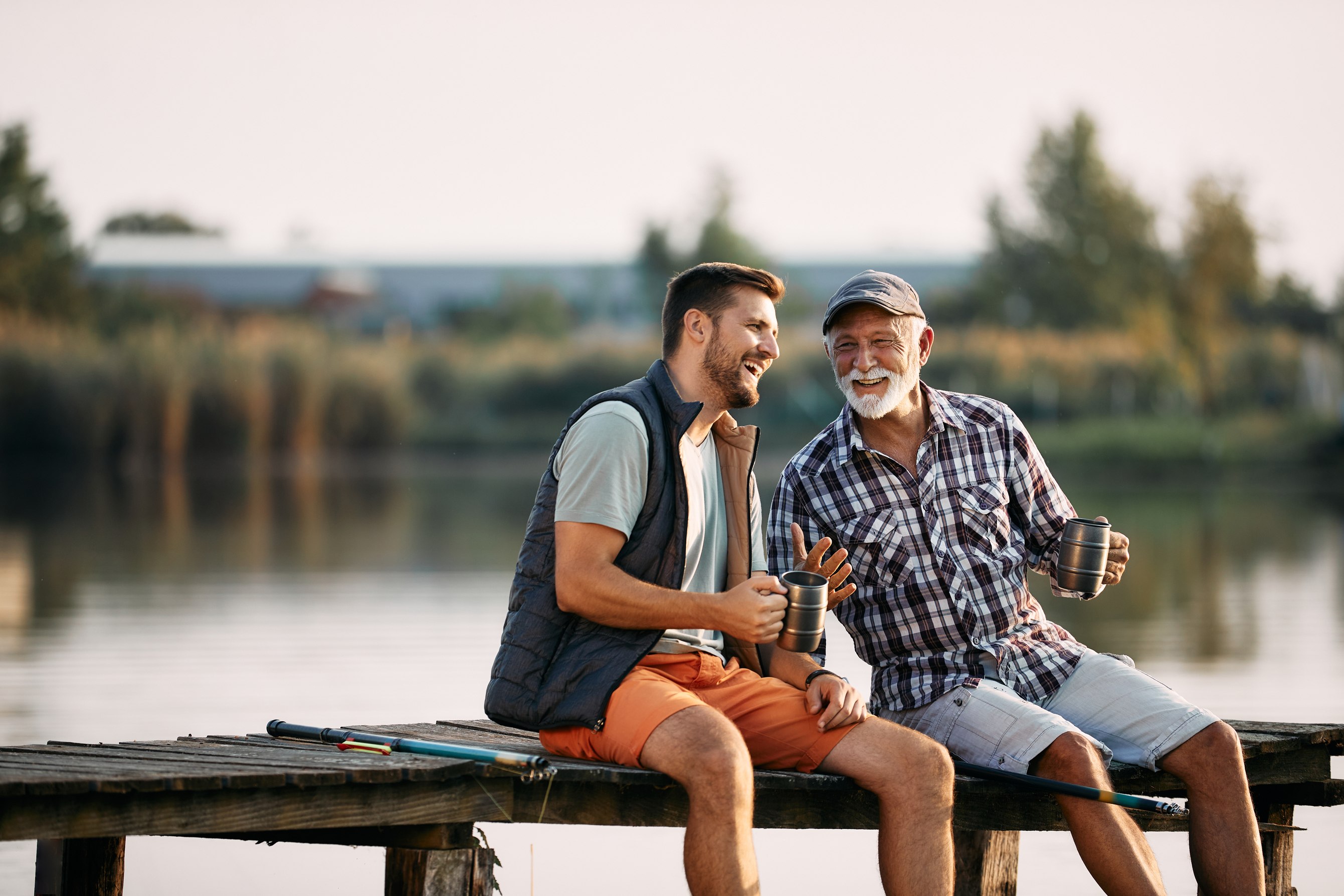 Two men laughing on wooden dock holding coffee mugs by lake