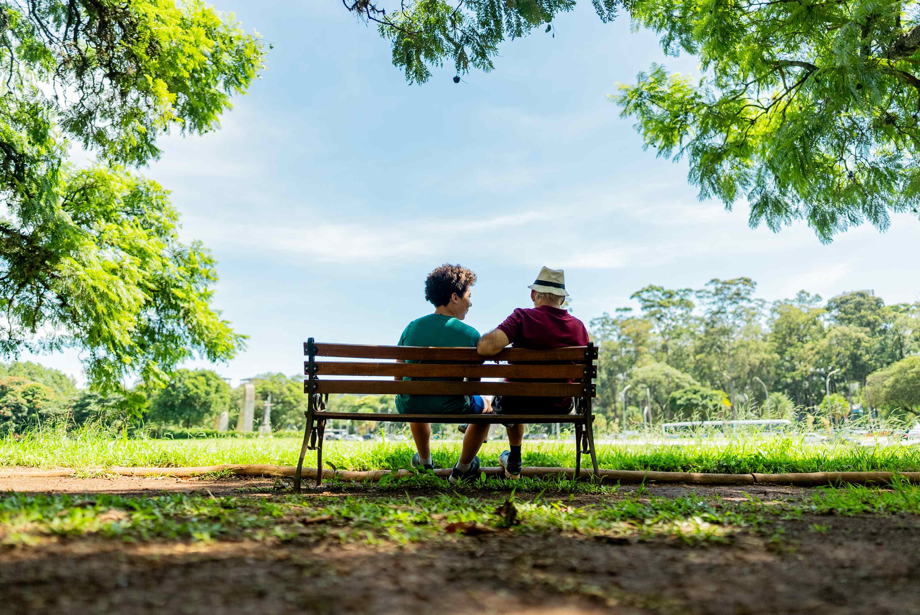 Two people sitting on park bench under trees on sunny day
