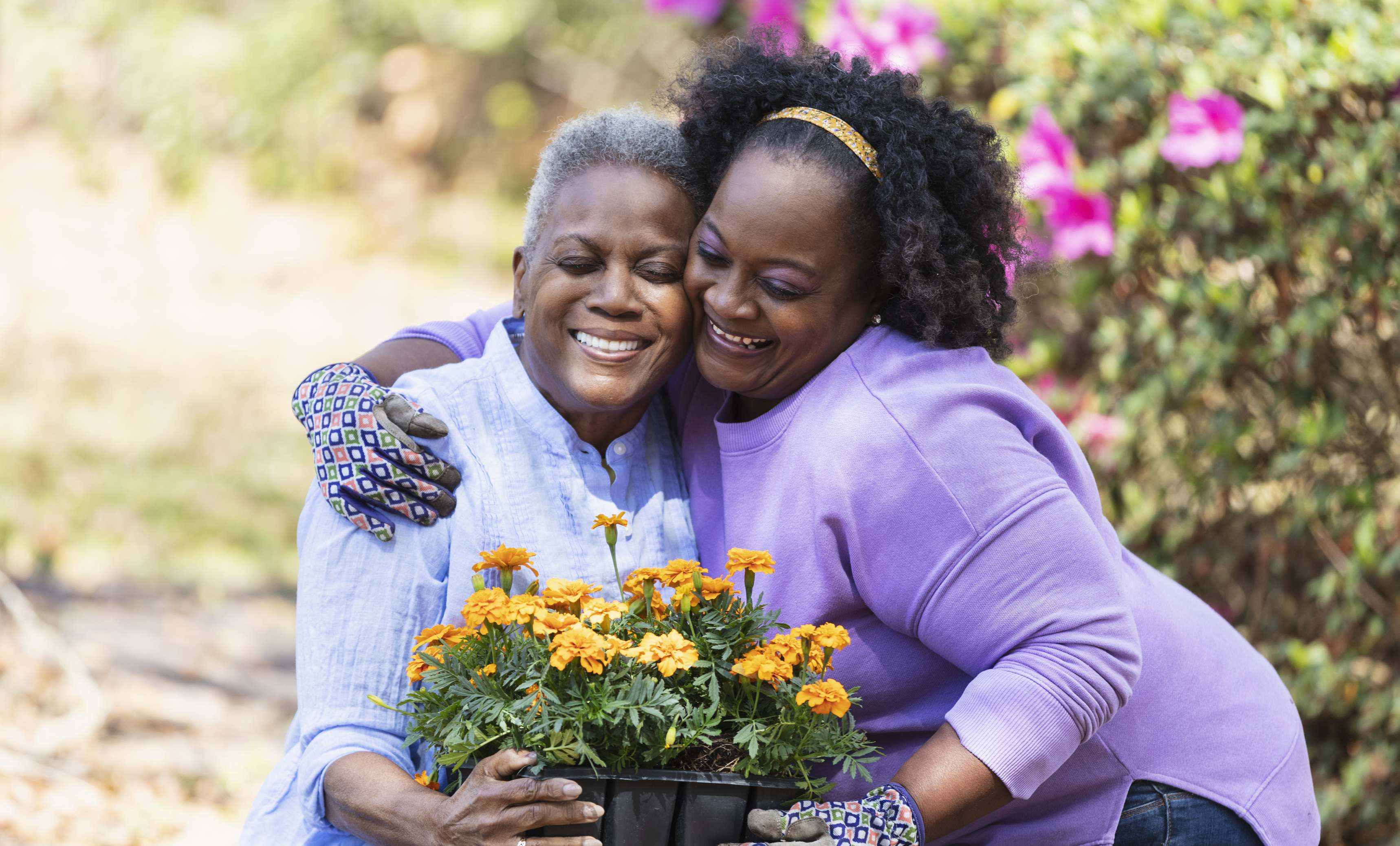 Two smiling women embracing while holding orange marigold flowers in garden