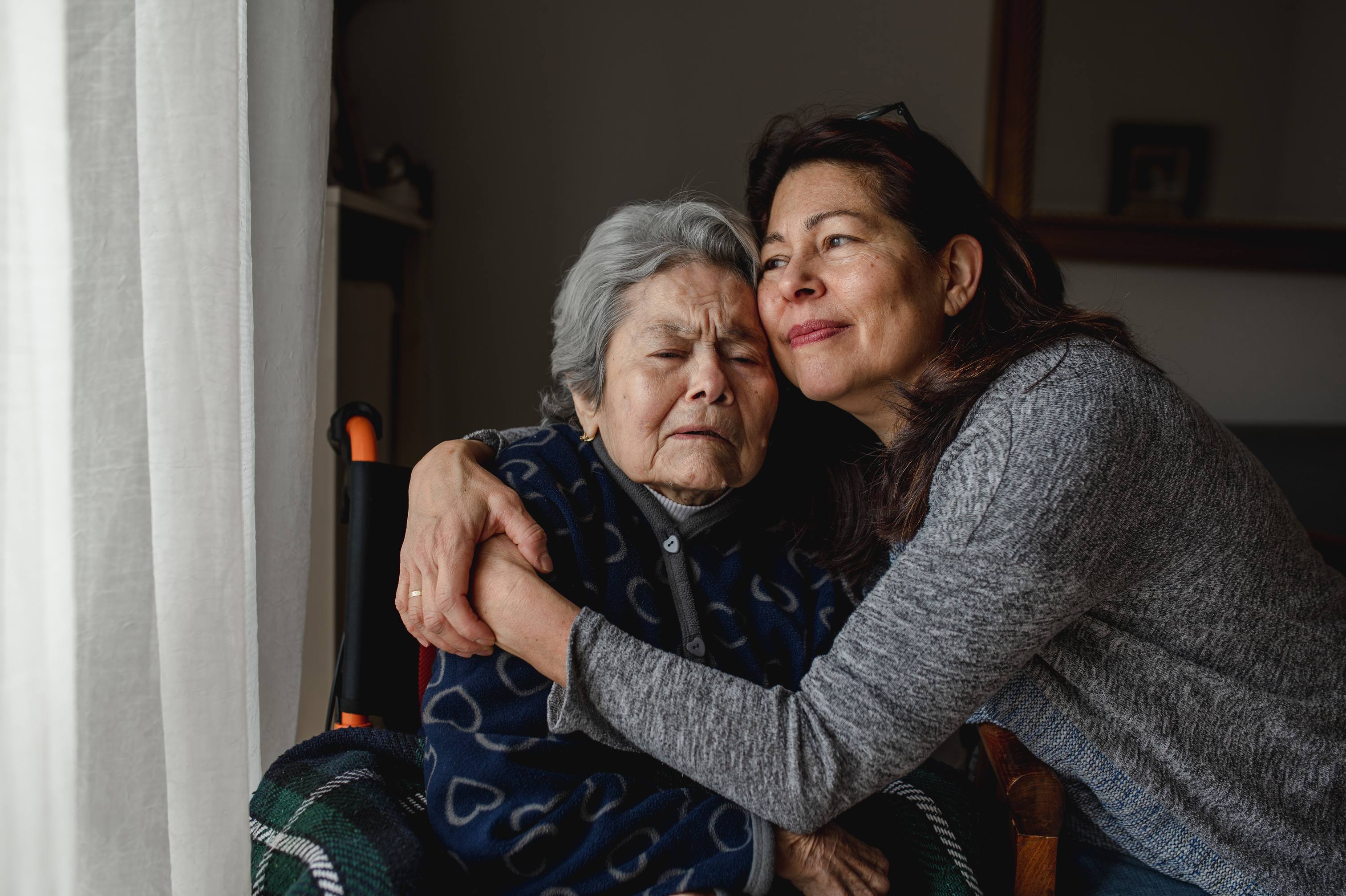 Two women embracing near a window, an elderly woman with gray hair being hugged by a younger woman, both showing emotional connection in soft natural light