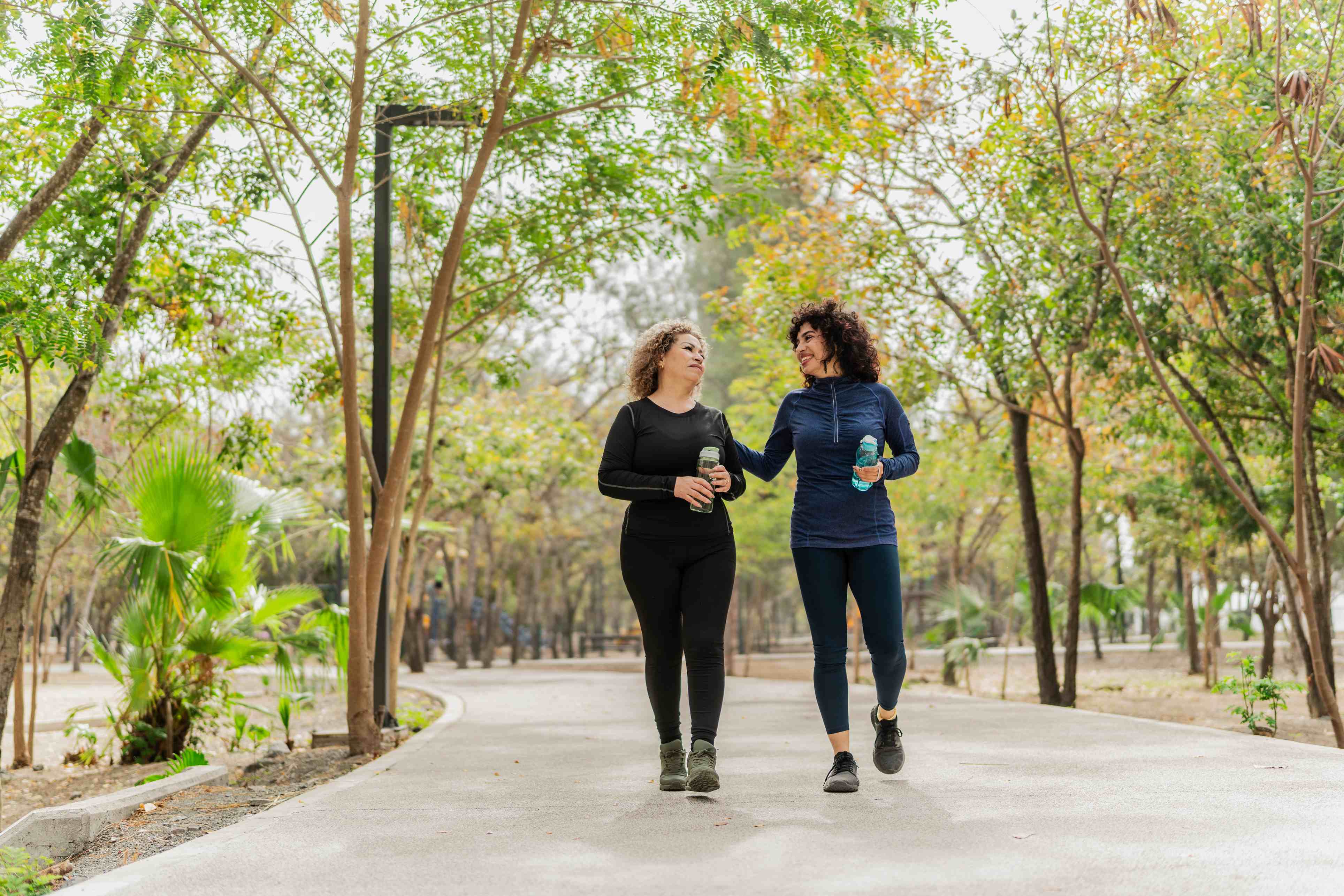 Two women walking together in park holding water bottles