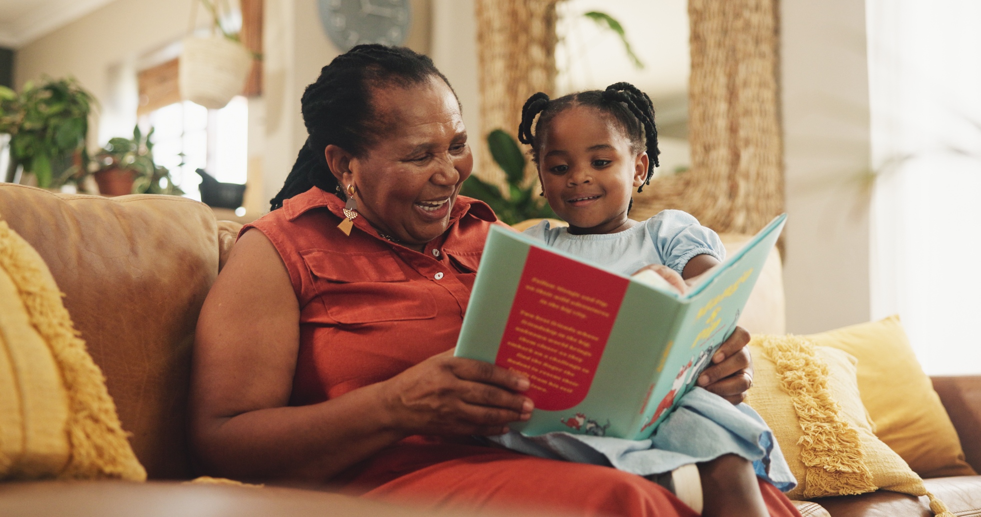 Woman in red shirt reading a book with young girl in blue dress on yellow couch