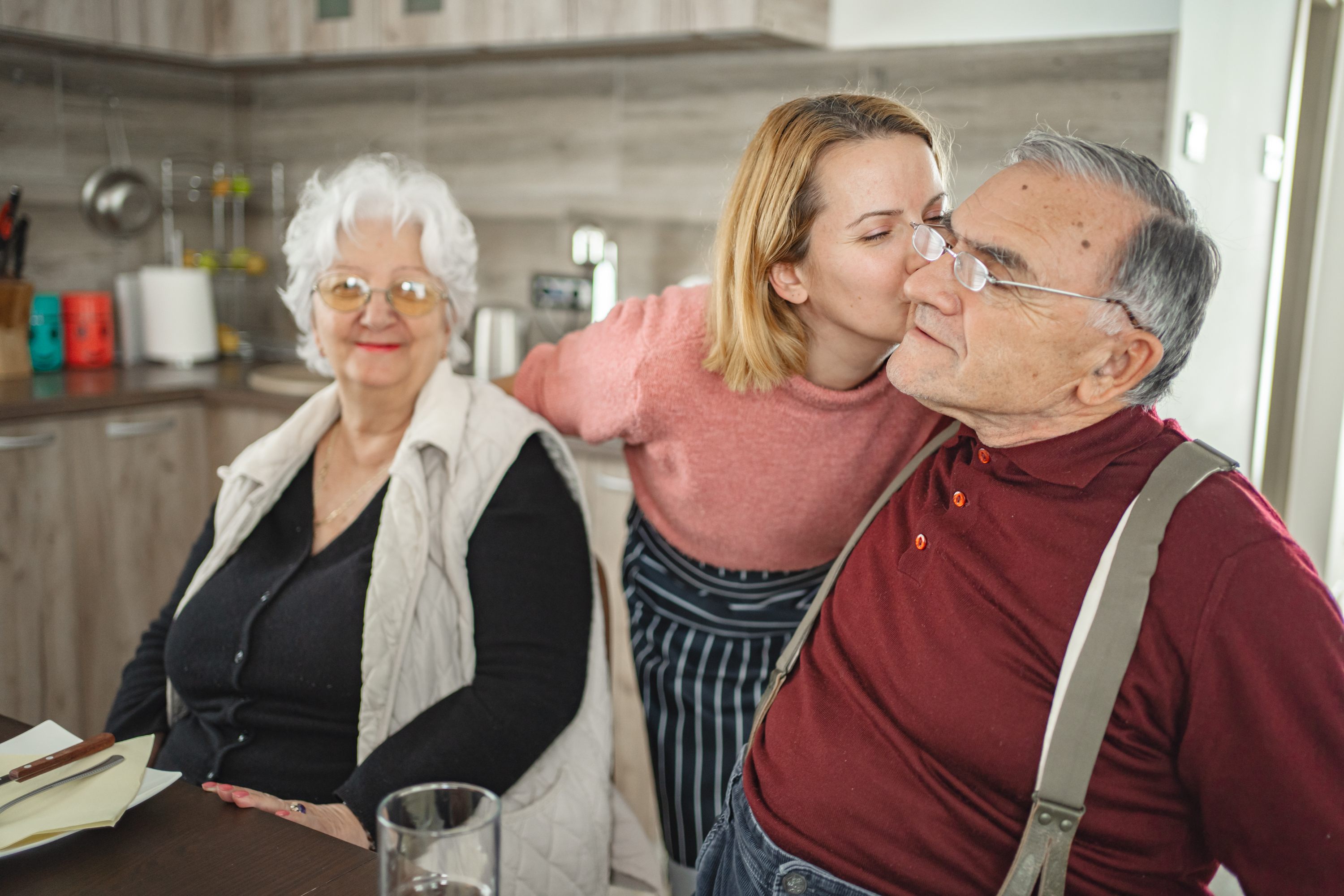 Woman kissing elderly man's cheek while elderly woman smiles in kitchen