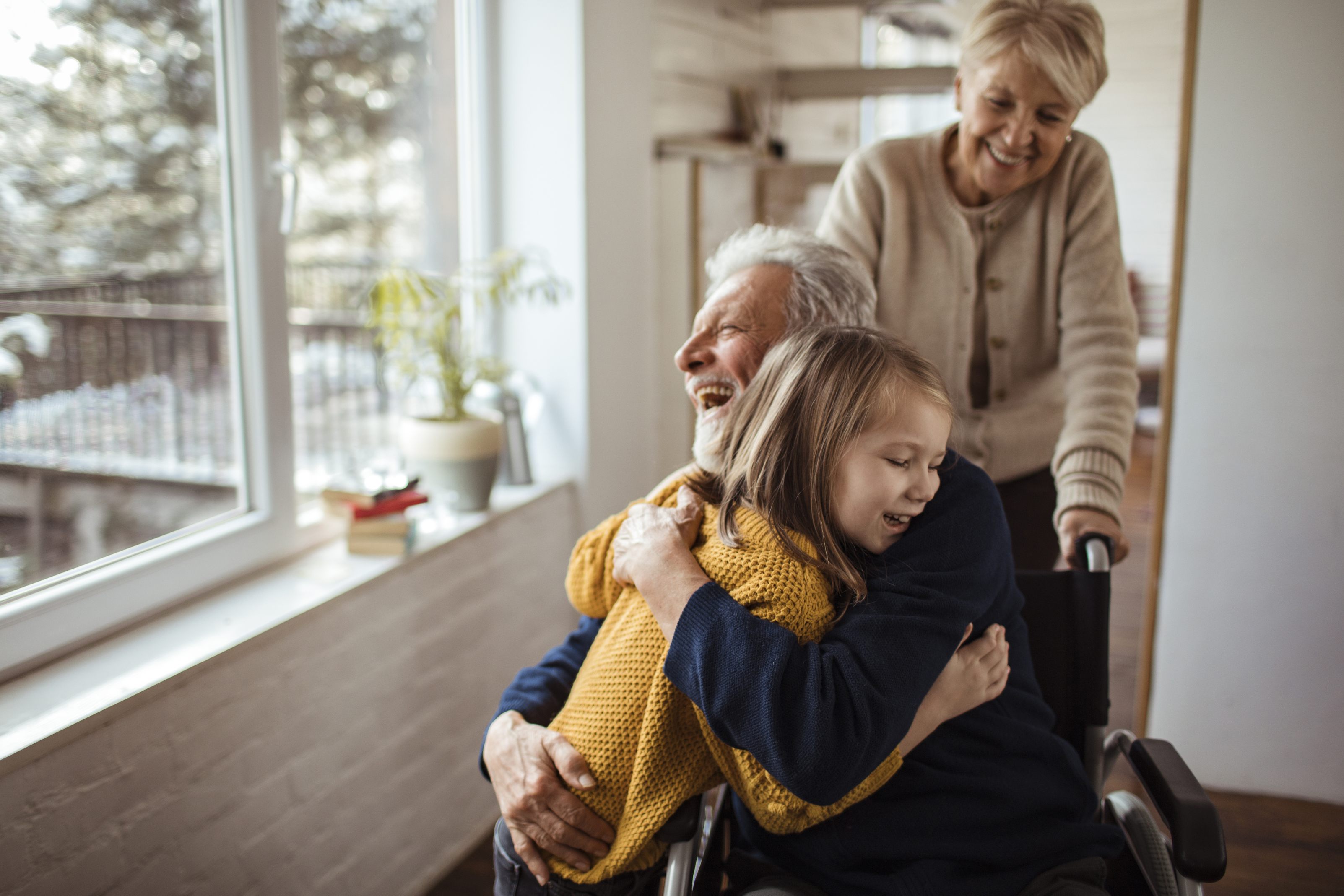 Young girl hugging elderly man in wheelchair while woman stands behind smiling